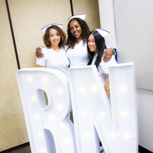 Three registered nursing graduates of Hudson County Community College, a training provider in the New Jersey Pay It Forward Program, pose in front of an "RN" sign at their pinning ceremony.